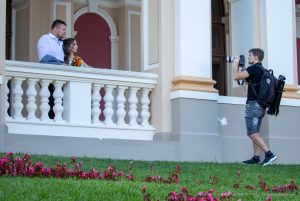 Фотография фотографа, снимающего пару молодоженов на фоне красивого здания. Видна татуировка на руке фотографа.