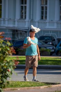 Фотография мужчины, идущего по улице с голубем, сидящим у него на голове. Яркий солнечный день, городская среда, необычный образ.