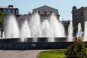 Фотография фонтана с брызгами воды на фоне зданий. Яркий солнечный день, голубое небо, зеленая трава. Идеально для вдохновения эскизов тату.