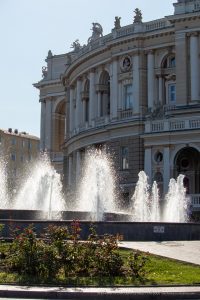 Фотография фонтана с брызгами воды на фоне красивого здания. На переднем плане видны красные розы.