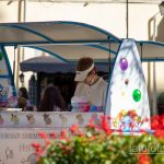 Фотография киоска с мороженым, украшенного яркими шариками и надписью 'ICE N' CO!'. Продавец в белой кепке готовит фруктовый лед.