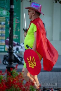 Фотография мужчины в необычном костюме: желтая футболка, красная накидка, розовая шляпа и щит с украинским гербом. В руке держит меч.