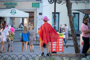 Фотография: человек в ярком красном плаще и розовой шляпе идет по улице, окруженный людьми и городскими пейзажами.
