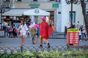 Фотография: человек в ярком красном плаще и розовой шляпе идет по улице, окруженный людьми и городскими пейзажами.