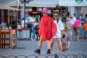 Фотография: человек в ярком красном плаще и розовой шляпе идет по улице, окруженный людьми и городскими пейзажами.