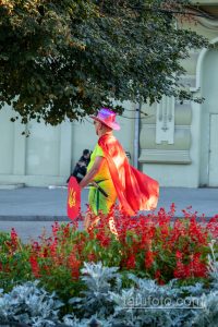 Фотография уличного артиста в красном плаще и розовой шляпе, держащего щит с украинским флагом на фоне здания.