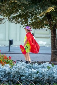 Фотография уличного артиста в красном плаще и розовой шляпе, держащего щит с украинским флагом возле дерева.