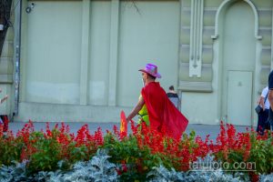Фотография уличного артиста в красном плаще и розовой шляпе, держащего щит с украинским флагом на фоне здания.