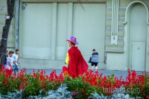 Фотография уличного артиста в красном плаще и розовой шляпе, стоящего на фоне здания и держащего щит.