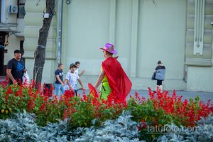 Фотография уличного артиста в красном плаще и розовой шляпе, держащего щит с украинским флагом.