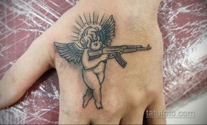 Фото тату на руке с изображением ангела с оружием. Черно-белый рисунок в стиле графика, с детально проработанными крыльями и автоматом.