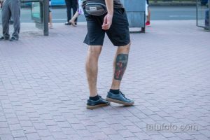 Фотография татуировки на голени мужчины, выполненной в ярких цветах. Рисунок виден сзади, когда мужчина стоит на улице.