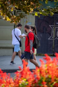 Фотография уличной татуировки: мужчина с татуировкой на руке идет по улице, мимо прохожих и цветочных клумб.