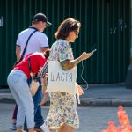 Фото девушки с татуировкой на руке и белой сумкой с надписью "Щастя" (Счастье). Девушка разговаривает по телефону на улице.