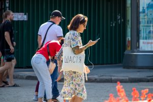 Фото девушки с татуировкой на руке и белой сумкой с надписью "Щастя" (Счастье). Девушка разговаривает по телефону на улице.