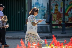 Фото девушки с татуировкой на руке и белой сумкой с надписью "Щастя" (Счастье). Девушка разговаривает по телефону на улице.