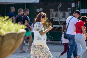 Фото девушки с татуировкой на руке и белой сумкой с надписью "Щастя" (Счастье). Девушка разговаривает по телефону на улице.