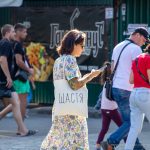 Фото девушки с татуировкой на руке и белой сумкой с надписью "Щастя" (Счастье). Девушка разговаривает по телефону на улице.