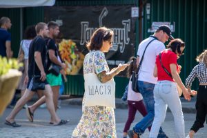 Фото девушки с татуировкой на руке и белой сумкой с надписью "Щастя" (Счастье). Девушка разговаривает по телефону на улице.