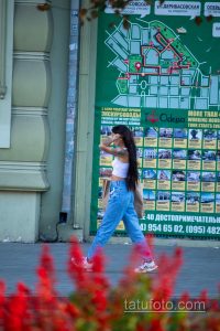 Фото девушки в джинсах и футболке с татуировкой на плече. Виден фрагмент рисунка татуировки, предположительно, цветочный мотив.