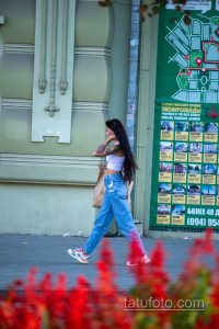 Фото девушки в джинсах и футболке с татуировкой на плече. Виден фрагмент рисунка татуировки, предположительно, цветочный мотив.