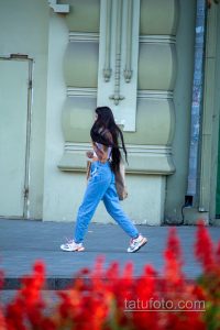Фото девушки в джинсах и футболке с татуировкой на плече. Виден фрагмент рисунка татуировки, предположительно, цветочный мотив.