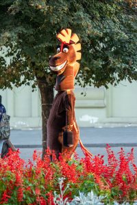 Фотография человека в костюме лошади, стоящего среди цветущих кустов. Костюм коричневого цвета с яркой гривой и хвостом.