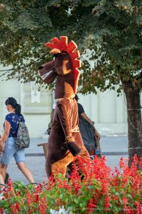 Фотография человека в костюме лошади, стоящего среди цветущих кустов. Костюм коричневого цвета с яркой гривой и хвостом.