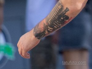 Фото крупным планом татуировки автомата Калашникова на предплечье мужчины. Детализированный рисунок татуировки с красными акцентами.