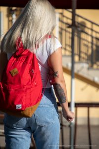 Фото тату: девушка с татуировкой птицы на руке, в белой футболке и светлых джинсах, с красным рюкзаком.
