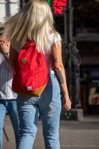 Фото тату на плече девушки, идущей по улице. Рисунок татуировки выполнен в ярких цветах, изображает пейзаж с элементами архитектуры.
