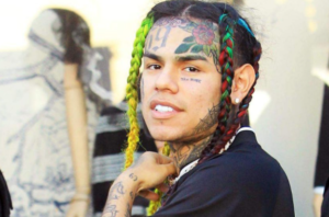 6ix9ine с татуировками на лице, включая цифры '69' на лбу и надпись 'THE WORLD', а также волосами, заплетенными в косички.