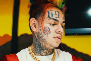 6ix9ine с татуировками на шее, лице и висках, включая цифры '69' на лбу и изображение черепа.