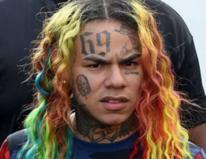 6ix9ine с разноцветными волосами и многочисленными татуировками на лице, включая цифры '69' на лбу.