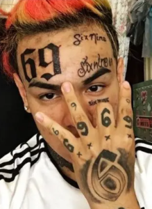 Крупный план лица 6ix9ine с многочисленными татуировками, включая цифры '69' на лбу и надписи на щеках.