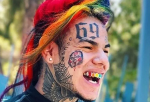 6ix9ine с татуировками на лице, шее и радужными волосами, демонстрирующий провокационный и яркий образ.