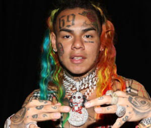 6ix9ine с радужными волосами, татуировками на лице, руках и массивным кулоном, демонстрирующий яркий и провокационный образ.