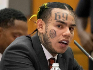 6ix9ine в официальном костюме с татуировками на лице и шее, демонстрирующий контраст между внешним видом и стилем.