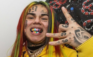 6ix9ine с татуировками на лице, руках и радужными волосами, демонстрирующий провокационный образ.