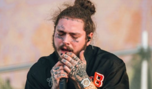 Фотография Post Malone во время выступления на сцене с многочисленными татуировками на лице, шее и руках. Видны надписи "Always" и "Blessed".