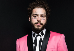 Фотография Post Malone в розовом пиджаке с многочисленными татуировками на лице, шее и руках. Видны надписи "Always" и "Blessed".