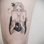 Фото тату на бедре с изображением девушки, читающей книгу под звездным небом. Детализированный рисунок татуировки в стиле графика с тонкими линиями.