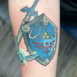 Фото тату: минималистичный рисунок щита и меча Линк из Zelda на руке, выполненный в простых линиях и цветах.
