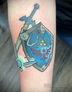 Фото тату: минималистичный рисунок щита и меча Линк из Zelda на руке, выполненный в простых линиях и цветах.