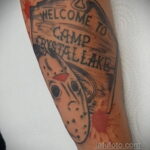 Фото тату маски Джейсона Вурхиза и надписи "Welcome to Camp Crystal Lake" на руке, выполненное в стиле олдскул с брызгами крови. Яркий рисунок татуировки.