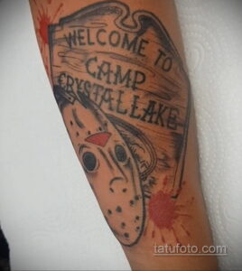 Фото тату маски Джейсона Вурхиза и надписи "Welcome to Camp Crystal Lake" на руке, выполненное в стиле олдскул с брызгами крови. Яркий рисунок татуировки.
