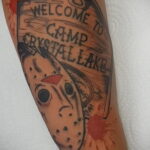 Фото тату маски Джейсона Вурхиза и надписи "Welcome to Camp Crystal Lake" на руке, выполненное в стиле олдскул с брызгами крови. Яркий рисунок татуировки.