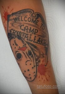 Фото тату маски Джейсона Вурхиза и надписи "Welcome to Camp Crystal Lake" на руке, выполненное в стиле олдскул с брызгами крови. Яркий рисунок татуировки.