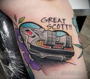 Фото тату на руке с изображением DeLorean, окруженного цветами, и надписью 'Great Scott!!'. Яркий и стилизованный рисунок.