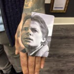 Фотография татуировки на руке с реалистичным изображением лица мужчины, разделенного на две части. Детализированный рисунок татуировки выполнен в черно-белых тонах.